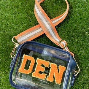 May include: A clear, rectangular crossbody bag with a navy blue trim and gold hardware. The bag features the word "DEN" in orange, fuzzy letters. The orange and silver striped strap is adjustable.