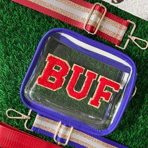 May include: Clear stadium bag with a blue trim and the letters "BUF" in red. The bag has a detachable strap with gold hardware and a red and white striped design. Additional straps with football patterns are also visible.