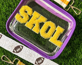 May include: A clear stadium-approved bag with a purple trim and the word "SKOL" in yellow, textured letters. The bag is surrounded by various straps, including a white strap with football designs and a yellow and white striped strap.