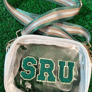 May include: A clear, white crossbody bag with a green and gold glitter strap. The bag features the letters "SRU" in green with a gold outline. The strap has green, gold, and silver stripes. The bag is on a green surface.