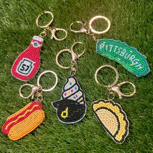 Puede incluir: Una colecci&oacute;n de llaveros con cuentas con dise&ntilde;os de comida y ciudad. Incluye una botella de ketchup roja, un perrito caliente, un cono de helado negro, un pierogi amarillo y un pepinillo verde con el texto "Pittsburgh". Todos los llaveros tienen herrajes dorados.