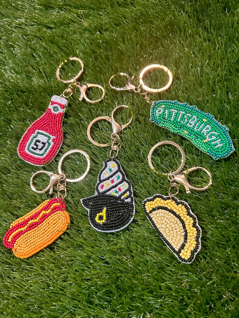 Puede incluir: Una colecci&oacute;n de llaveros con cuentas con dise&ntilde;os de comida y ciudad. Incluye una botella de ketchup roja, un perrito caliente, un cono de helado negro, un pierogi amarillo y un pepinillo verde con el texto "Pittsburgh". Todos los llaveros tienen herrajes dorados.