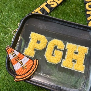 Puede incluir: Bolso bandolera transparente con ribete negro, una correa amarilla y negra y las letras "PGH" en amarillo. Un charm de cono de tr&aacute;fico naranja y blanco con cuentas est&aacute; adjunto a la bolsa. La bolsa est&aacute; sobre una superficie verde.