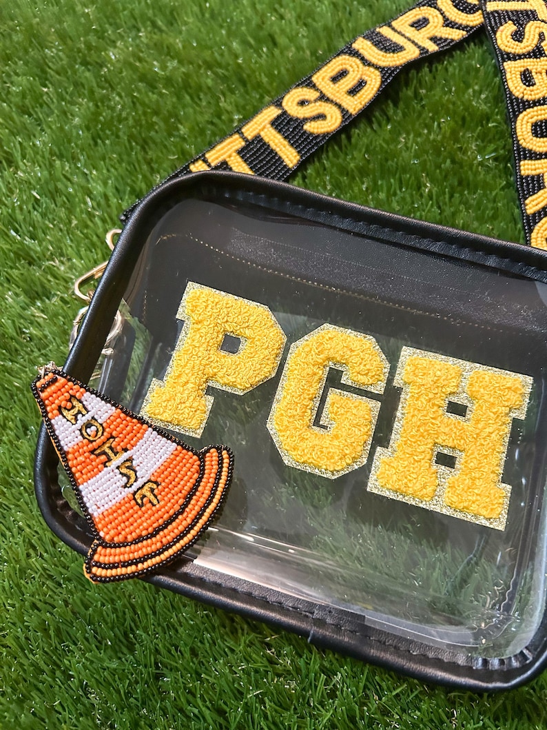 Puede incluir: Bolso bandolera transparente con ribete negro, una correa amarilla y negra y las letras "PGH" en amarillo. Un charm de cono de tr&aacute;fico naranja y blanco con cuentas est&aacute; adjunto a la bolsa. La bolsa est&aacute; sobre una superficie verde.