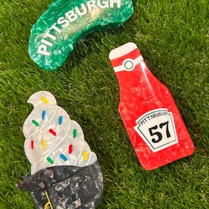 May include: Three Pittsburgh-themed accessories on green turf. A green, translucent, pickle-shaped item with "PITTSBURGH" in white. A red ketchup bottle with "PITTSBURGH 57". A cupcake-shaped item with white frosting and colorful sprinkles.