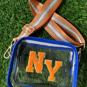 May include: Clear crossbody bag with a blue trim and an orange and white striped strap. The bag features the orange letters "NY" on the front. The strap is adjustable and has gold-colored hardware.