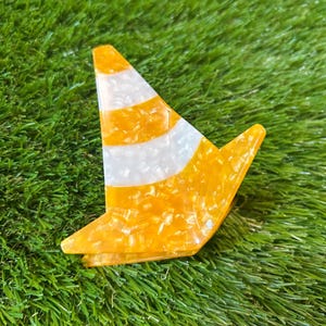 May include: A hair claw clip shaped like a traffic cone, featuring a marbled orange and white pattern. The accessory has a pointed top and a wide base, constructed from a glossy, translucent acrylic material with a distinct speckled texture.