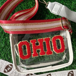 May include: Clear stadium-approved crossbody bag with the word "OHIO" in red and gold lettering. The bag has a red, white, and gold striped strap and a white strap with football-shaped embellishments. The background is green.