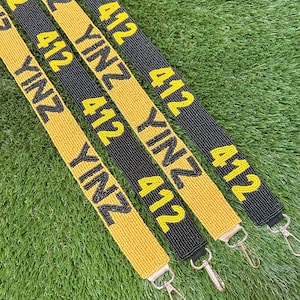 May include: Four beaded straps with the text "YINZ 412" in yellow on black and yellow backgrounds. Each strap has a metal clasp at the end. The straps are lying on a bed of green grass.