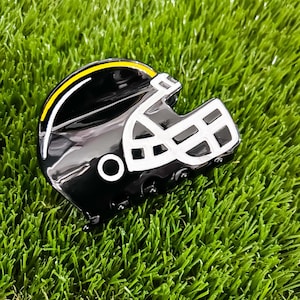 Peut inclure: Une pince à cheveux noire en forme de casque de football américain. Le casque présente une finition noire brillante, une grille de protection blanche, une bande jaune sur le dessus et un petit cercle blanc sur le côté. Le mécanisme est intégré.