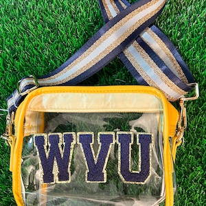 May include: A clear, yellow crossbody bag with the letters "WVU" in navy blue and gold. The bag has a striped strap in navy, gold, and light blue. The bag is on a green surface.