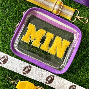 May include: Clear stadium-approved bag with purple trim and gold hardware. The bag features the letters "MIN" in gold, with a yellow and white striped strap, a solid purple strap, and a white strap with football designs.