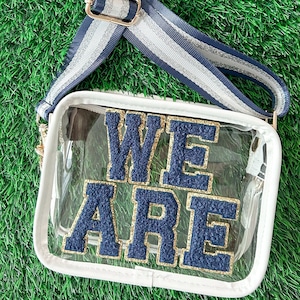 May include: A clear, rectangular crossbody bag with the words "WE ARE" in navy blue and gold lettering. The bag has a white trim, a gold chain, and a striped navy and silver strap. The bag is on a green surface.