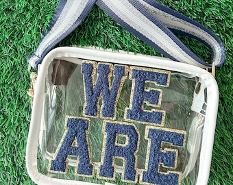 Puede incluir: Un bolso cruzado rectangular transparente con las palabras "WE ARE" en letras azul marino y doradas. El bolso tiene un ribete blanco, una cadena dorada y una correa a rayas azul marino y plateada. El bolso está sobre una superficie verde.