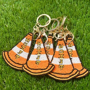 Puede incluir: Varios llaveros de cuentas con forma de conos de tr&aacute;fico naranjas y blancos con la palabra "HOIST" en letras amarillas. Los llaveros tienen anillos y cierres dorados, sobre un fondo de c&eacute;sped artificial verde.