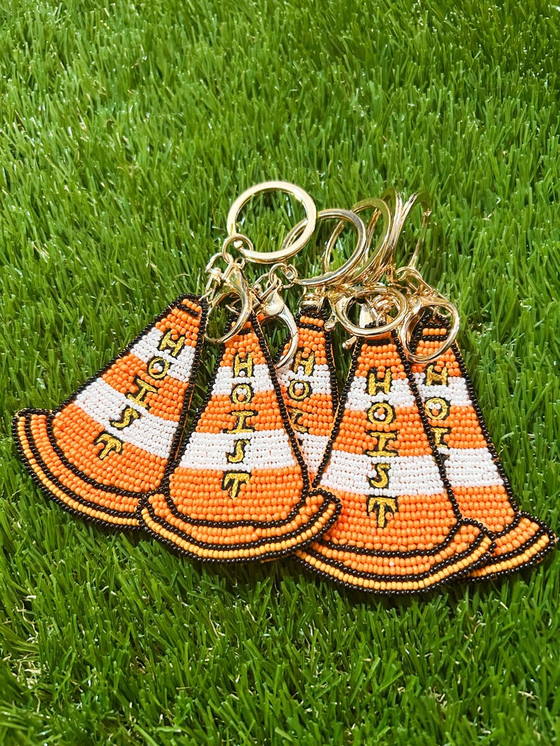 Puede incluir: Varios llaveros de cuentas con forma de conos de tr&aacute;fico naranjas y blancos con la palabra "HOIST" en letras amarillas. Los llaveros tienen anillos y cierres dorados, sobre un fondo de c&eacute;sped artificial verde.