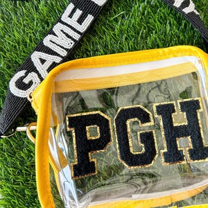 May include: Clear stadium bag with yellow trim and the letters "PGH" in black and gold. The bag has a black and white beaded strap that says "GAME". The bag is on a bed of green grass.