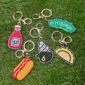 May include: A collection of beaded keychains featuring food and city-themed designs. Includes a ketchup bottle, hot dog, ice cream cone, pierogi, and a keychain that says "Pittsburgh". Each keychain has a gold-colored keyring.