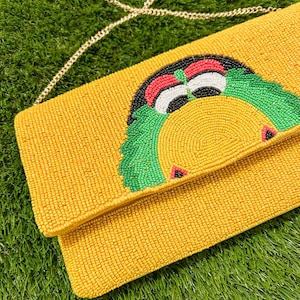 May include: Yellow beaded clutch with a gold chain strap. The clutch features a colorful beaded design of a bird with green, red, black, and white accents. The clutch is on a green surface.