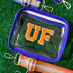 May include: A clear stadium-approved bag with a blue border and a white and blue striped zipper. The bag features the orange letters "UF". Two detachable straps, one blue and one orange, with silver and gold glitter stripes, are also included.