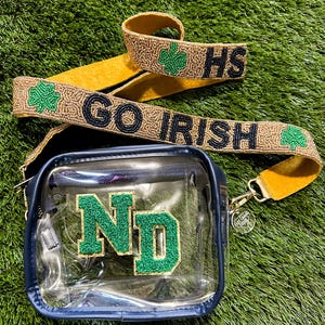 May include: A clear, navy blue crossbody bag with a green and gold "ND" patch. The bag has a gold beaded strap with "GO IRISH" and "HS" in black beads, along with green shamrock accents. The bag is on a green surface.