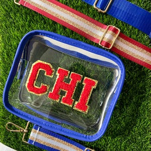 May include: Clear crossbody bag with a blue trim and the letters "CHI" in red and gold. The bag is shown with several colorful straps, including red with gold glitter, blue, and white with gold and blue stripes. The straps have gold-colored metal hardware.