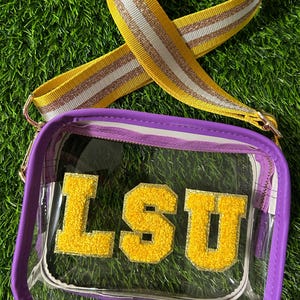 May include: Clear crossbody bag with a purple frame and a yellow and white striped strap. The bag features the letters "LSU" in yellow, textured lettering. The bag is on a green surface.
