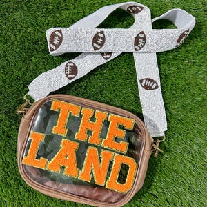 May include: A brown crossbody bag with a clear front panel displaying the text "THE LAND" in orange block letters. The bag has a white beaded strap with brown football designs. Gold-colored hardware.
