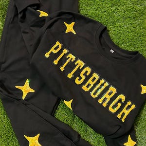 May include: Black sweatshirt and sweatpants set. The sweatshirt features the word "PITTSBURGH" in gold sequined letters. Gold sequined stars are on the sweatshirt and sweatpants. The set is displayed on a green surface.