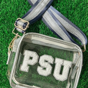 May include: Clear crossbody bag with silver trim and a navy and silver striped strap. The bag features the letters "PSU" in white, with a silver glitter effect. The bag is on a green surface.