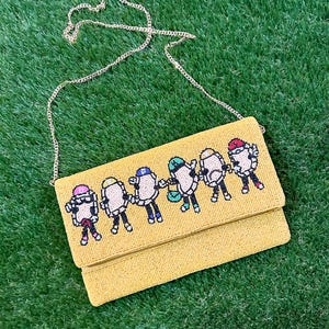 May include: Yellow beaded clutch with a gold chain strap. The clutch features a pixelated design of six figures holding hands, each wearing a different colored hat. The background is green.