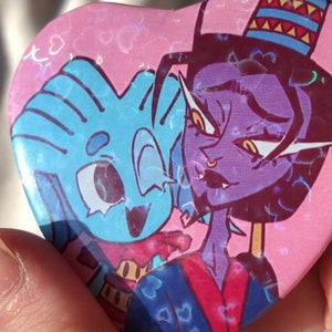 May include: Heart-shaped button with a cartoon illustration of two characters. One character is blue with a striped head, the other is purple with a hat. The background is pink with heart-shaped holographic accents.