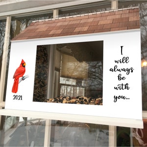 May include: A white bird feeder with a red cardinal perched on a branch. The feeder has a window with birdseed inside. The feeder has a roof and the text "I will always be with you..." on the right side. The year 2021 is printed below the cardinal.