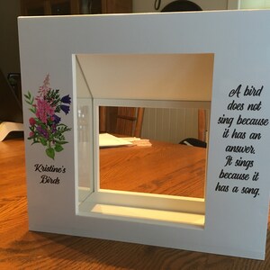 May include: White wooden birdhouse with a glass front and a floral design on the side. The birdhouse has a quote on the right side that reads: "A bird does not sing because it has an answer. It sings because it has a song."