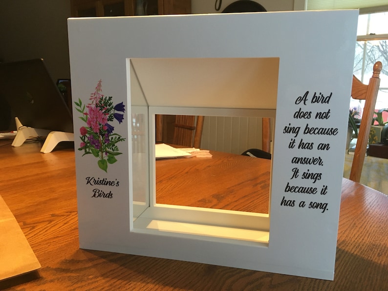 May include: White wooden birdhouse with a glass front and a floral design on the side. The birdhouse has a quote on the right side that reads: "A bird does not sing because it has an answer. It sings because it has a song."