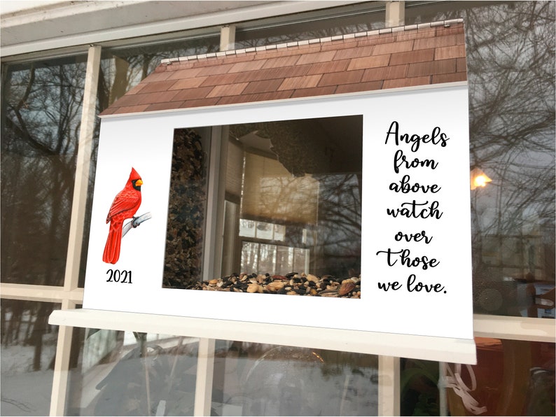 May include: A white bird feeder with a red cardinal perched on a branch. The feeder has a window and a roof. The text "Angels from above watch over those we love." is printed on the right side of the feeder. The year 2021 is printed on the left side of the feeder.