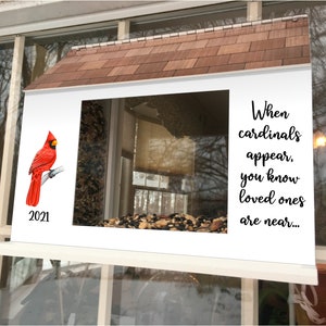 May include: A white bird feeder with a red cardinal perched on a branch. The feeder has a wooden roof and a clear window. The text "When cardinals appear, you know loved ones are near..." is printed on the feeder. The year 2021 is printed on the bottom left corner of the feeder.