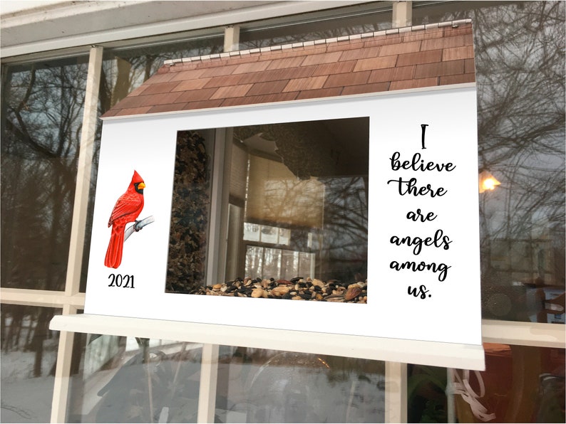 May include: A white bird feeder with a red cardinal perched on a branch. The feeder has a window with a view of the outside. The feeder has a roof and the text "I believe there are angels among us." on the right side. The year 2021 is printed on the left side of the feeder.