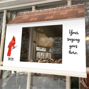 May include: A white bird feeder with a red cardinal perched on a branch. The feeder has a brown shingled roof and a clear window. The text "Your saying goes here." is printed on the right side of the feeder. The year 2021 is printed below the cardinal.