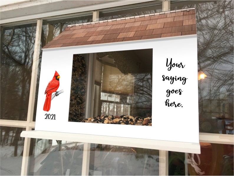 May include: A white bird feeder with a red cardinal perched on a branch. The feeder has a brown shingled roof and a clear window. The text "Your saying goes here." is printed on the right side of the feeder. The year 2021 is printed below the cardinal.