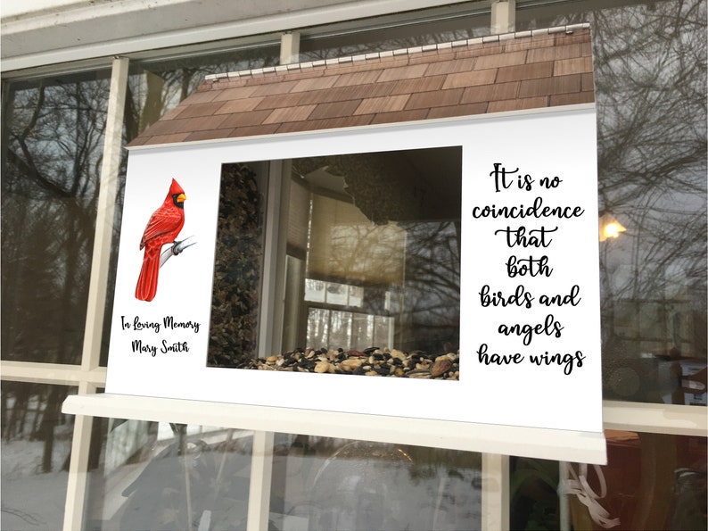 May include: A white bird feeder with a red cardinal perched on a branch. The feeder has a clear window and is filled with birdseed. The feeder is mounted on a window and has a sign that reads "It is no coincidence that both birds and angels have wings."  The sign also includes the text "In loving memory Mary Smith".
