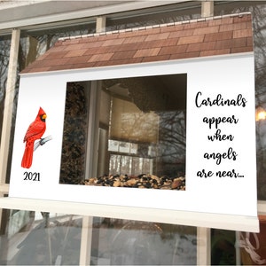May include: A white bird feeder with a red cardinal perched on a branch. The feeder has a brown roof and a clear window. The text "Cardinals appear when angels are near..." is printed on the feeder. The year 2021 is printed on the bottom left corner of the feeder.