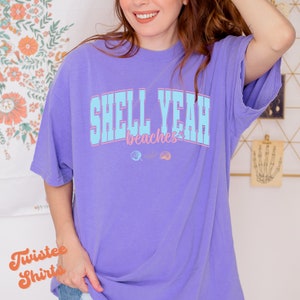 May include: A purple t-shirt with the text "Shell Yeah Beaches" in blue and pink lettering. There are two small shell graphics below the text.
