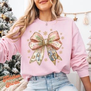 May include: Pink Christmas sweatshirt with a gold glitter bow and a nativity scene inside the bow. The bow has a gold glitter trim and a green garland.