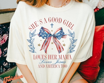 May include: A white t-shirt with the text "She's a good girl loves her mama loves Jesus and America too" in red, blue, and gold lettering. A watercolor illustration of a red, white, and blue bow with a gold cross above it is centered on the shirt.