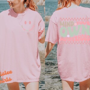 May include: Pink t-shirt with a smiley face on the front and the text "Mind Your Own Motherhood" on the back. The text is in a checkered pattern with pink, green, and white colours.