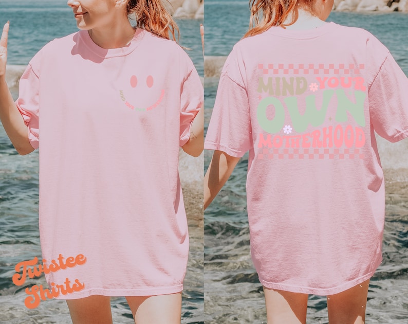 May include: Pink t-shirt with a smiley face on the front and the text "Mind Your Own Motherhood" on the back. The text is in a checkered pattern with pink, green, and white colours.