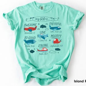 May include: A mint green short-sleeve t-shirt featuring a graphic print of airplanes and helicopters with clouds and stars. The text reads: my GOD is... AWESOME Nehemiah 1:5, FAITHFUL Lam. 3:22-23, LOVE 1 John 4:8, SO GOOD Psalm 34:8, MIGHTY Zephaniah 3:17, STRONG Psalm 28:7.