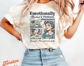Puede incluir: Camiseta color crema con el texto "Emotionally Attached to Fictional Characters". El gráfico incluye ilustraciones de una copa de vino, libros y una mujer. También se incluye el texto "Wines and coffees help!" y "Therapy? Nah, just more books".
