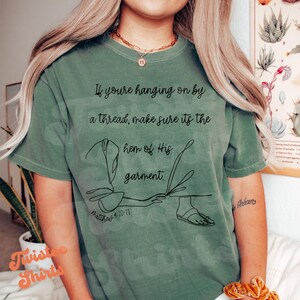 May include: Sage green t-shirt with the text "If you're hanging on by a thread, make sure it's the hem of His garment." A line drawing of a person's feet and garment is below the text. The text "Matthew 9:20-22" is also on the shirt.
